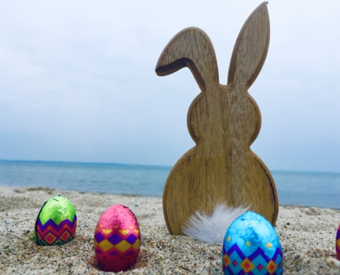 Osterhase aus Holz, bunte Eier drumherum im Sand, Meer Im Hintergrund